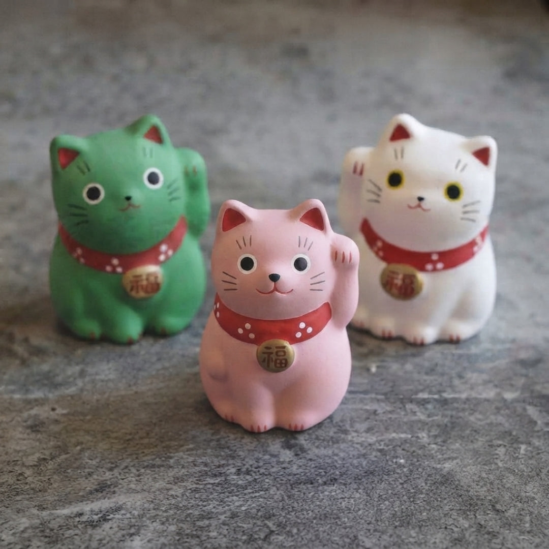 Trois statuettes Maneki Neko rose, verte et blanche présentées ensemble dans une ambiance japonaise.