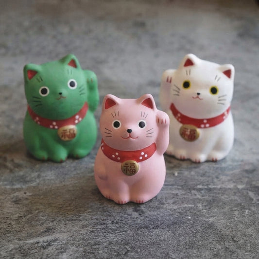 Trois statuettes Maneki Neko rose, verte et blanche présentées ensemble dans une ambiance japonaise.