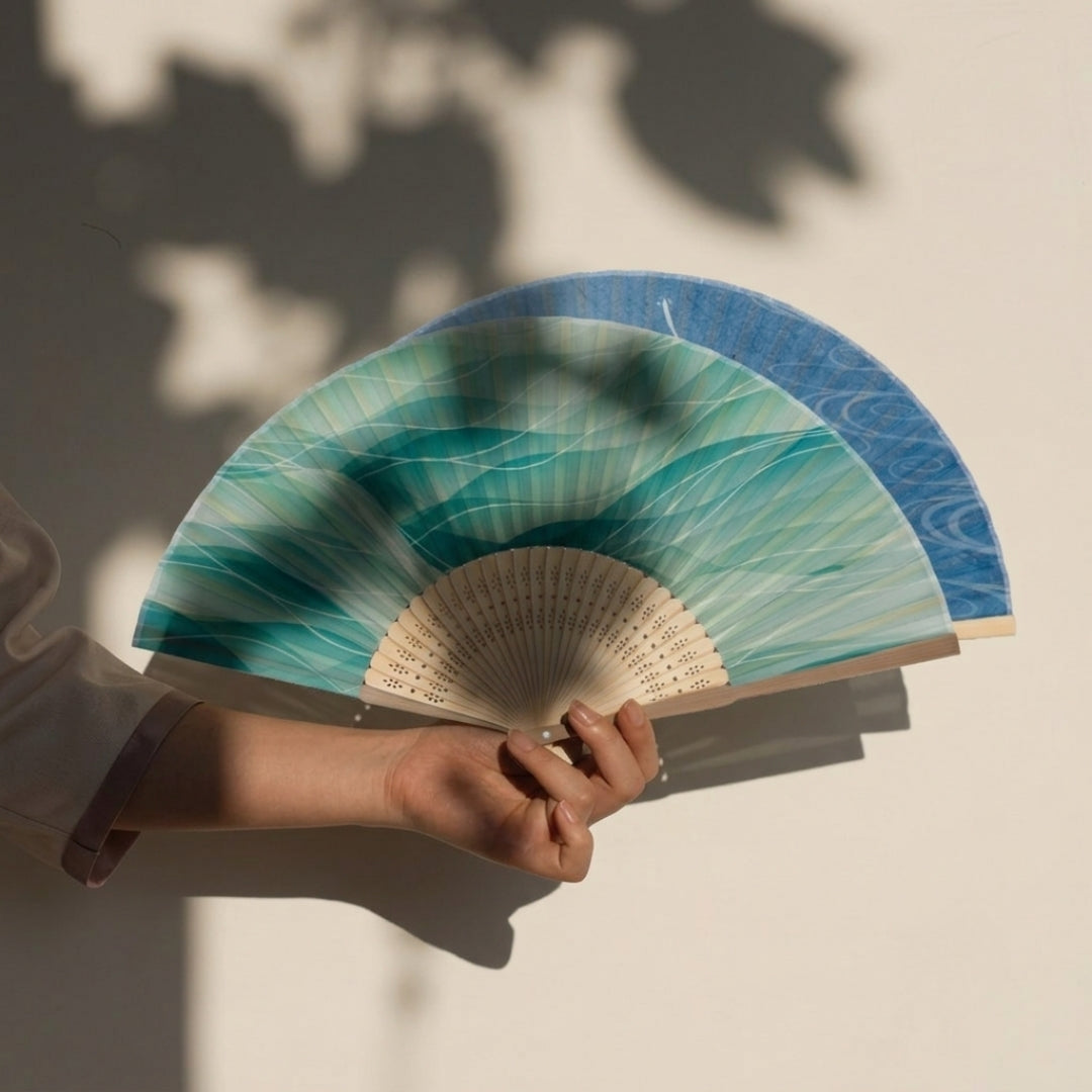 Main tenant les deux modèles d'éventails japonais en bambou, motifs vagues turquoise et poissons bleus.