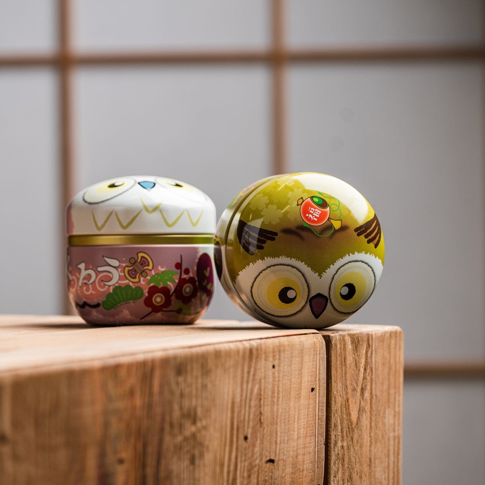 Boîtes à thé Yugen lab SUZOKU en fer blanc, forme ronde de hibou, coloris jaune et rose, posées sur un bloc de bois.