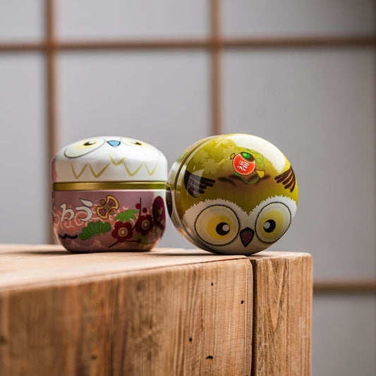 Boîtes à thé Yugen lab SUZOKU en fer blanc, forme ronde de hibou, coloris jaune et rose, posées sur un bloc de bois.