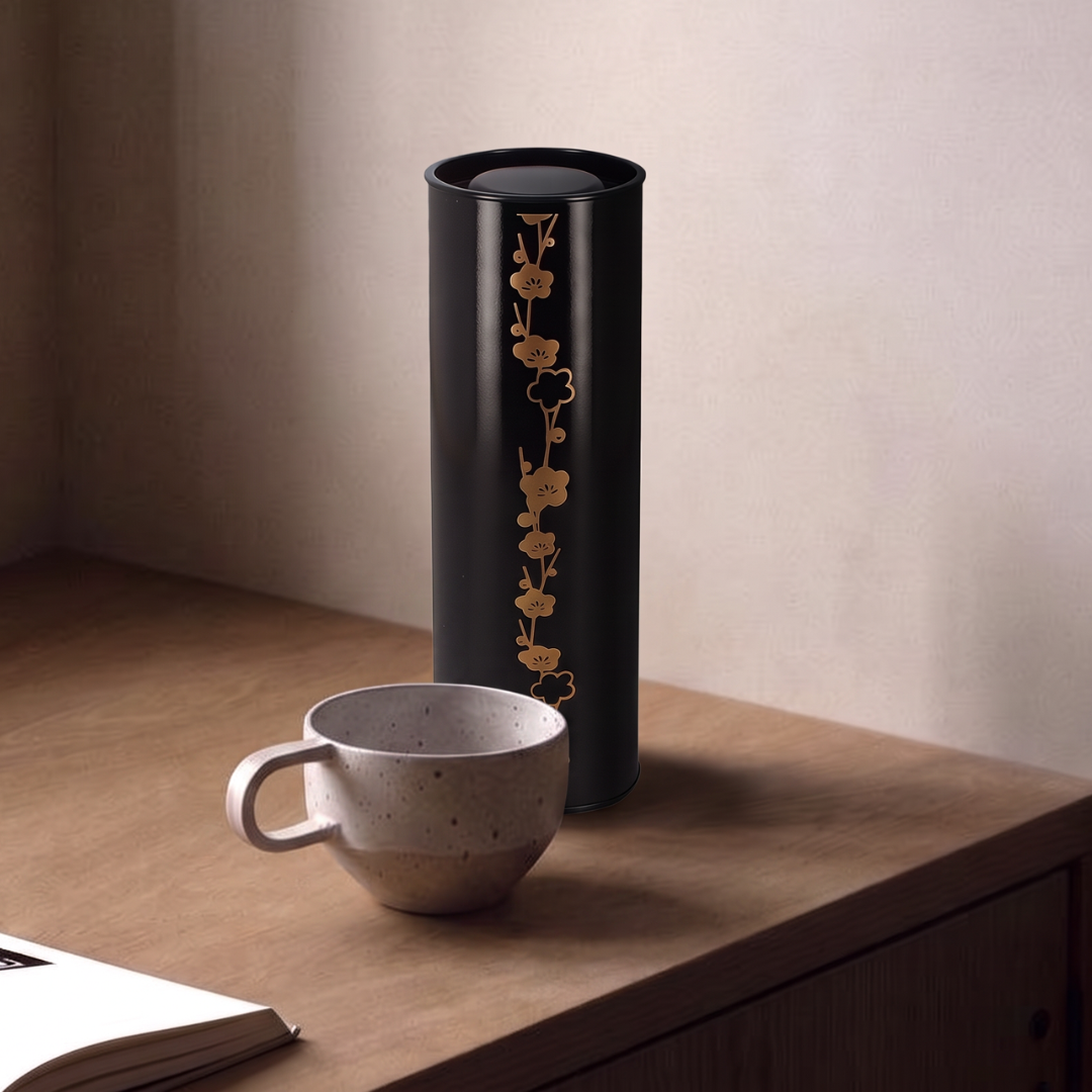 Boîte à thé Yugen lab noire et dorée en fer blanc avec motif de fleurs de prunier, posée sur une table en bois.