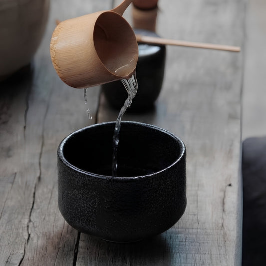 Bol à matcha Yugen lab en céramique noire texturée recevant de l'eau chaude versée depuis une louche en bambou