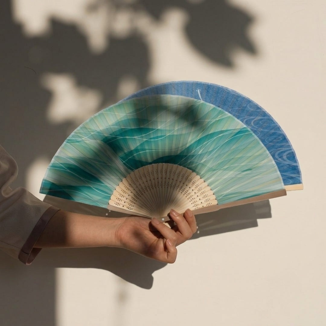 Éventail japonais Yugen lab en bambou et polyester avec motif vagues turquoise, tenu à la main dans une lumière douce.