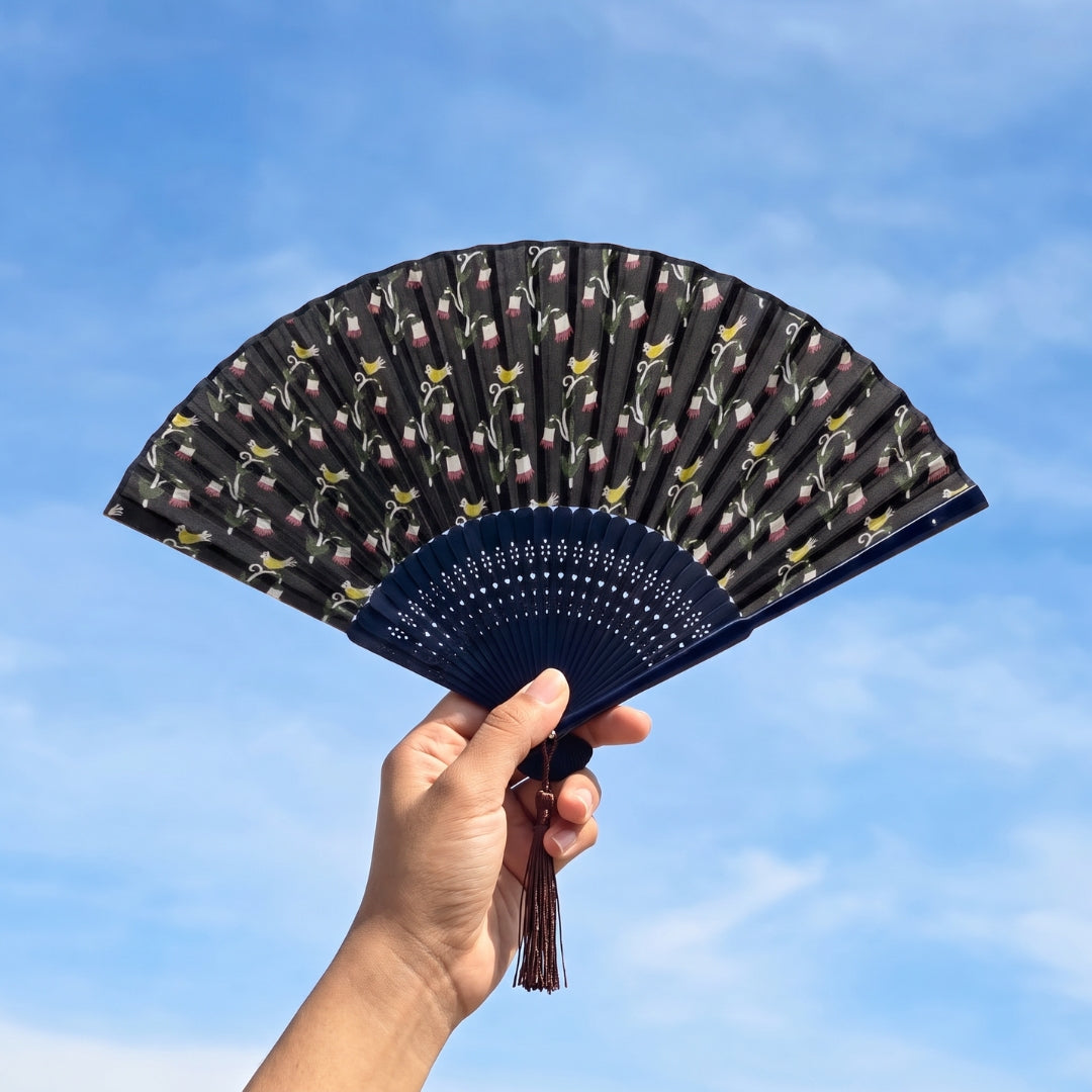 Éventail japonais Yugen lab en tissu bleu foncé avec motifs de fleurs et oiseaux jaunes, tenu devant un ciel bleu.