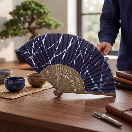 Éventail Yugen lab en tissu bleu marine et beige avec motif abstrait, posé sur une table en bois dans un intérieur.