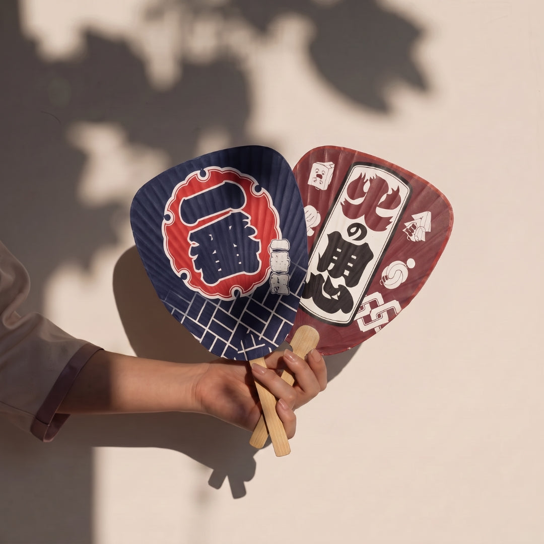 Main tenant deux éventails japonais uchiwa Yugen lab en bambou avec motifs graphiques traditionnels Ichiban et Hi no Yojin