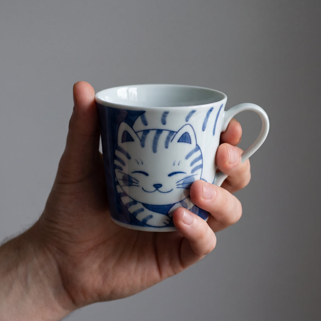 Main tenant un mug japonais Yugen lab en céramique blanche avec un motif de chat tigré bleu, finition brillante.