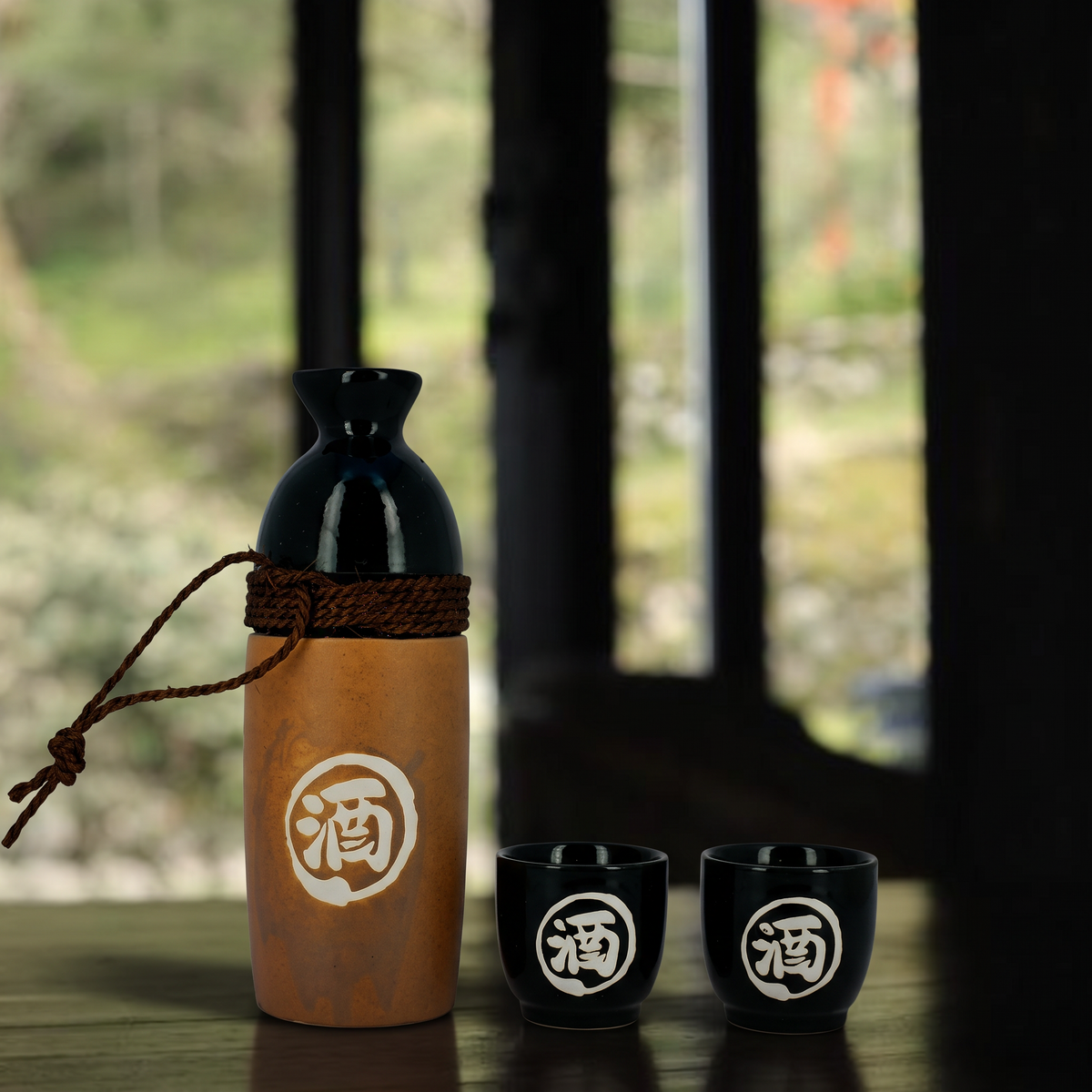 Service à saké japonais brun et noir à cordon, carafe et deux coupes devant une fenêtre ouverte sur un jardin